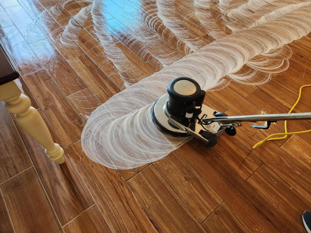 A floor buffer machine is being used to scrub and clean a wood-look tile floor, leaving behind visible soap patterns.