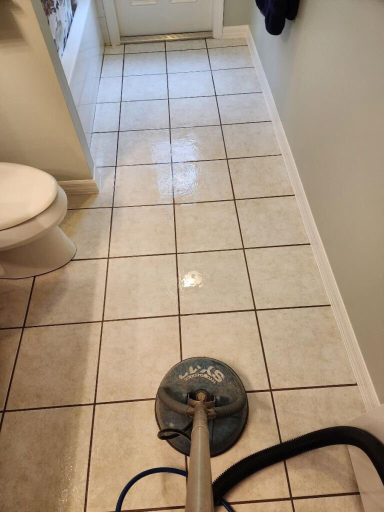 The image shows a tile floor in a bathroom being cleaned with a professional floor cleaning machine.