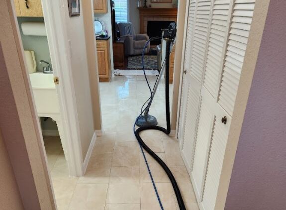 The image shows a hallway with a vacuum cleaner in the center, leading into a living room area with a fireplace and kitchen visible in the background.