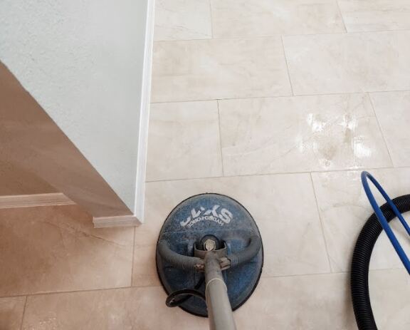 The image shows a close-up view of a tile floor being cleaned using a professional floor cleaning machine with hoses attached.