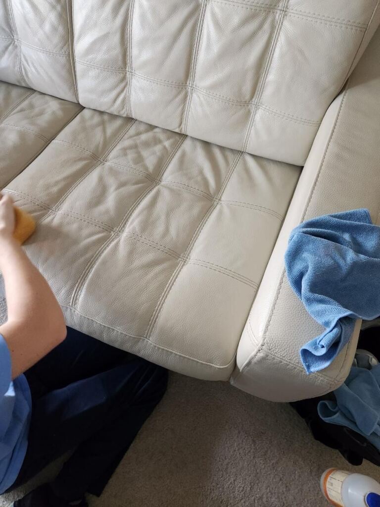 The image shows a person cleaning a leather couch using a sponge, with a blue microfiber cloth placed nearby on the couch.