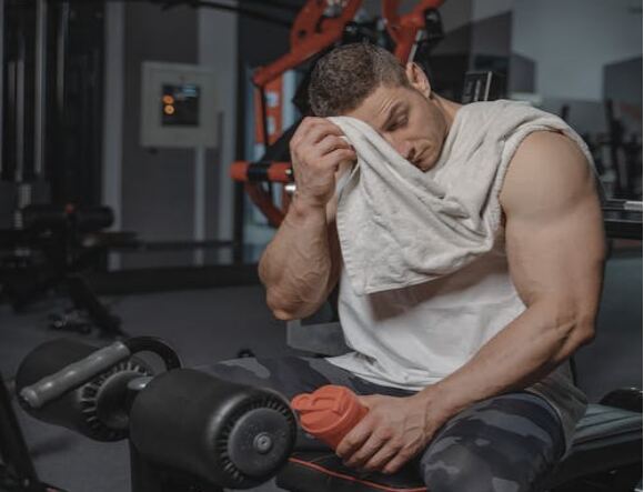 The image shows a man taking a break in the gym, wiping his forehead with a towel after an intense workout.