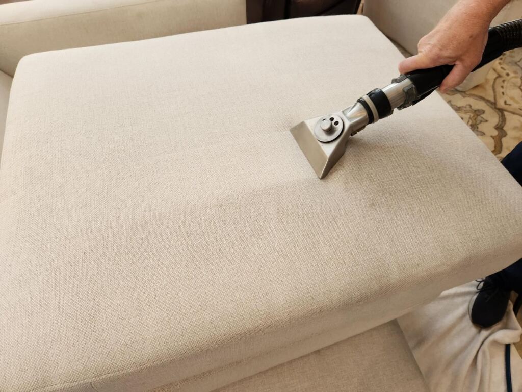 A person is cleaning a beige fabric couch cushion using a professional upholstery cleaning tool.